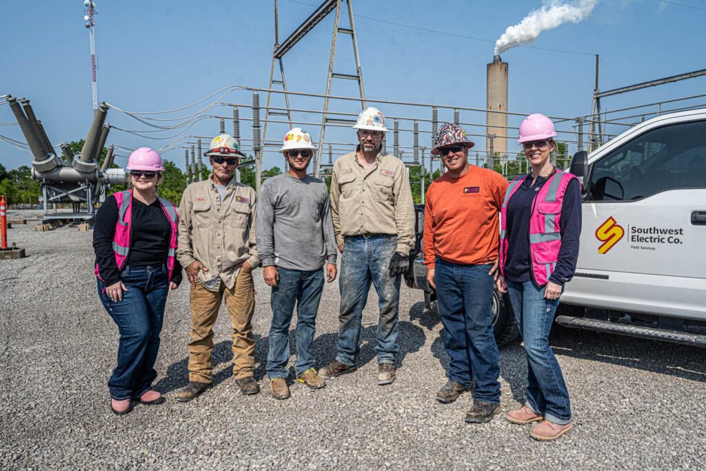 A group of Southwest Electric Co. field service technicians and team members wearing hard hats and safety gear, standing in a substation site with industrial equipment in the background.