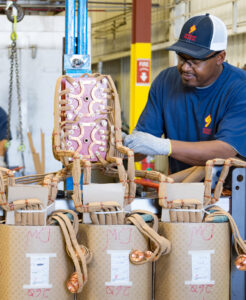 Technician assembling transformer windings with copper components in an industrial facility, using safety gloves and equipment for precision.